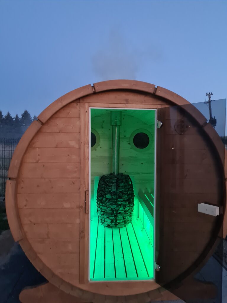 Drewniana sauna beczkowa z zielonym oświetleniem wewnątrz, widoczna po zmroku.